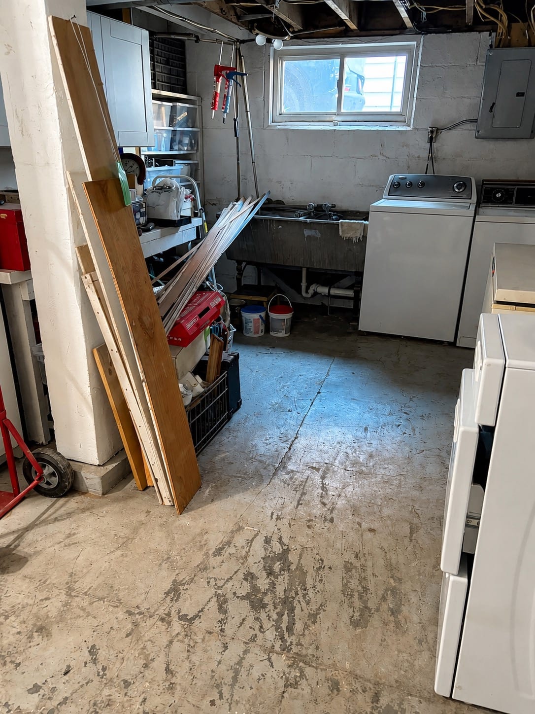 Basement before work: bare stained concrete floor with lumber, tools and buckets stacked against the wall beside a utility sink