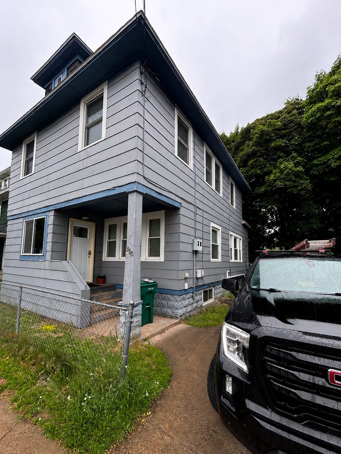 Two-story house before siding work: weathered gray siding with peeling blue porch trim and a bare dirt yard