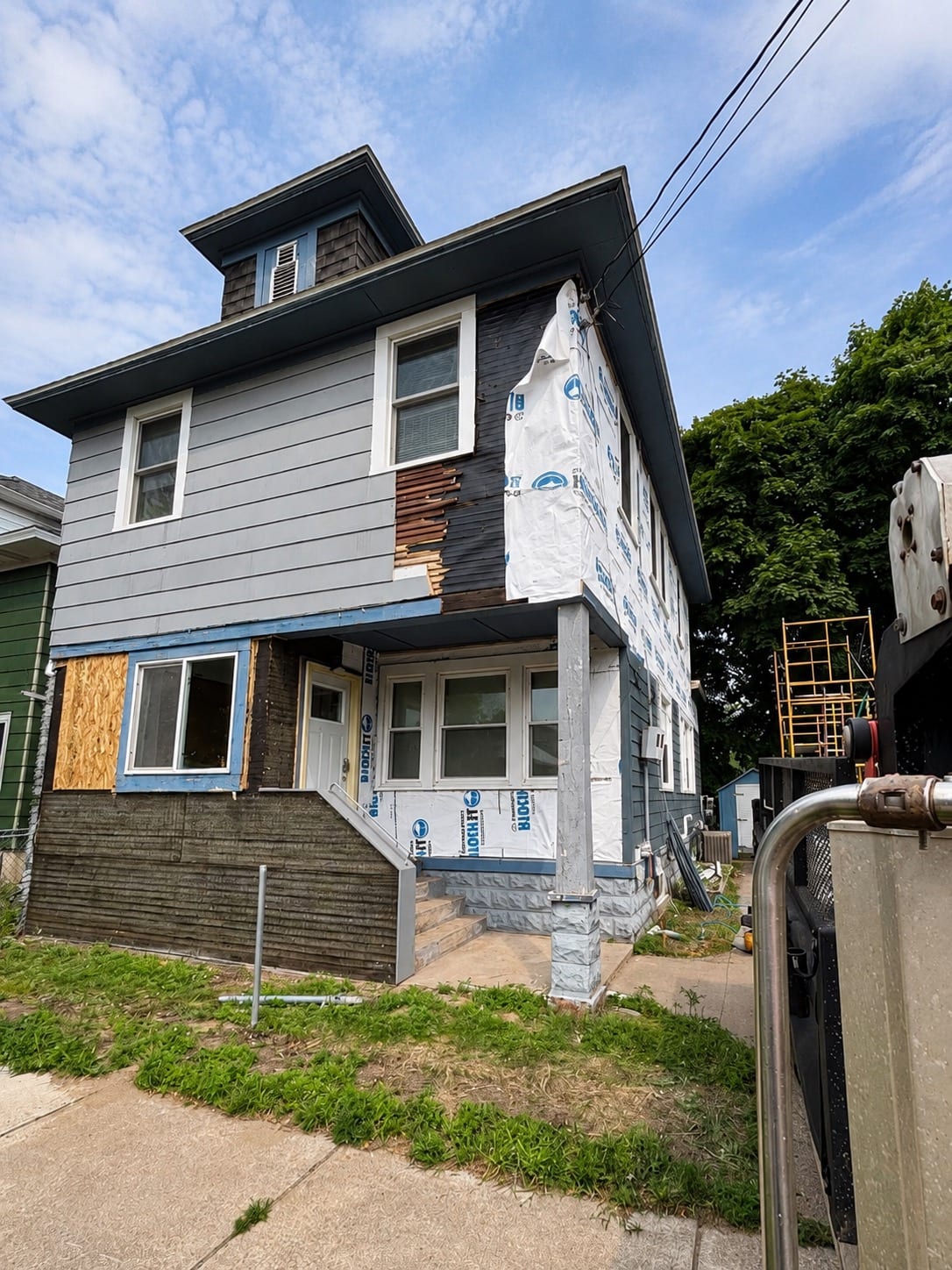 The same house mid-project: old siding stripped to reveal rotted boards and fresh house wrap, with one window boarded and scaffolding staged alongside