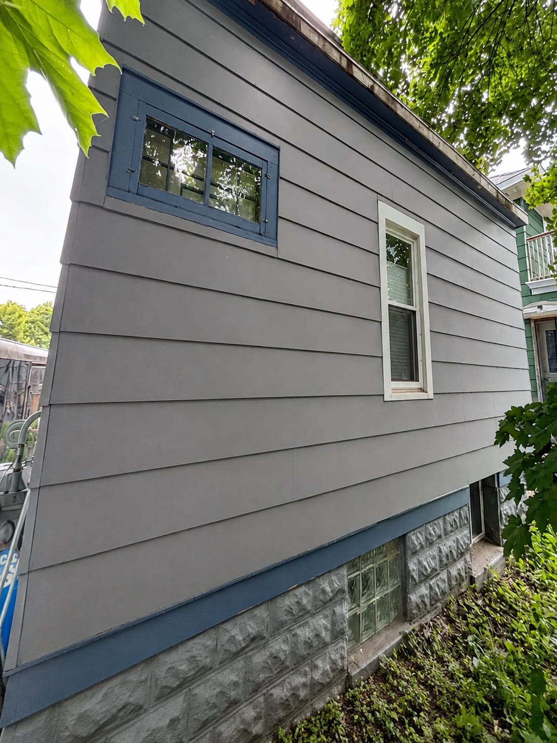 Finished gray lap siding meeting a stone-faced foundation, with blue-trimmed basement windows and a glass block window below the siding line